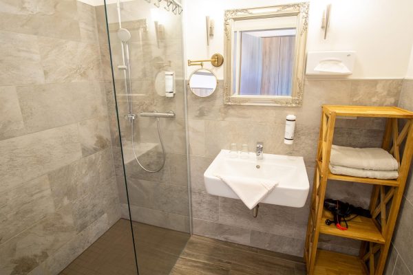 Modernes Hotelbadezimmer mit hellgrauen Fliesen und einem Holzschrank. Einem Föhn und bodenebener Dusche.