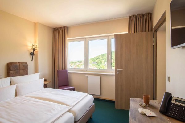 Das Bett vom Doppelzimmer mit Verbindungstür und die großen Fenster mit Gardinen und Ausblick zur Stadt Wernigerode.
